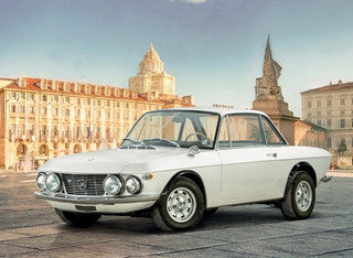 A vintage white Lancia Fulvia  coupe is parked outdoors in a city square with historic buildings and towers in the background.