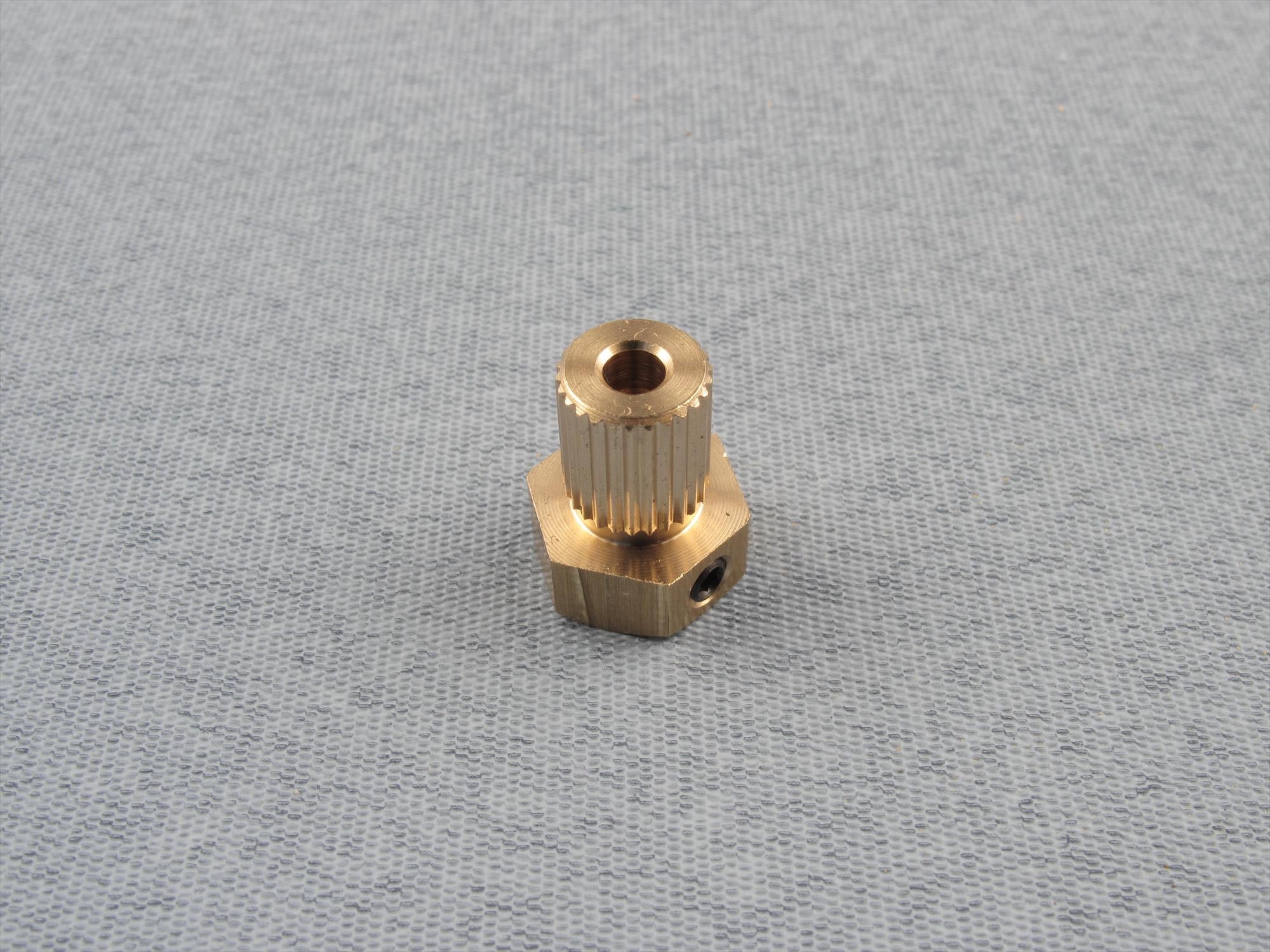 A small brass hexagonal threaded component with a knurled top section, photographed on a light gray textured fabric background.