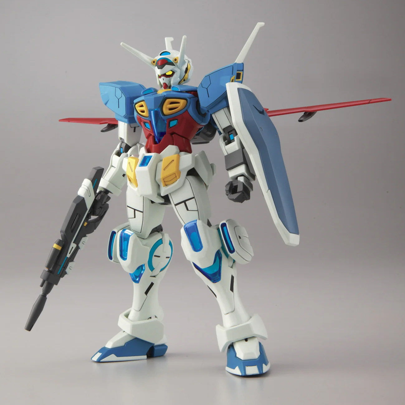 A detailed plastic model of a Gundam mecha figure, standing in a dynamic pose and holding a rifle in one hand and a shield with red wings in the other, set against a neutral gray studio background.